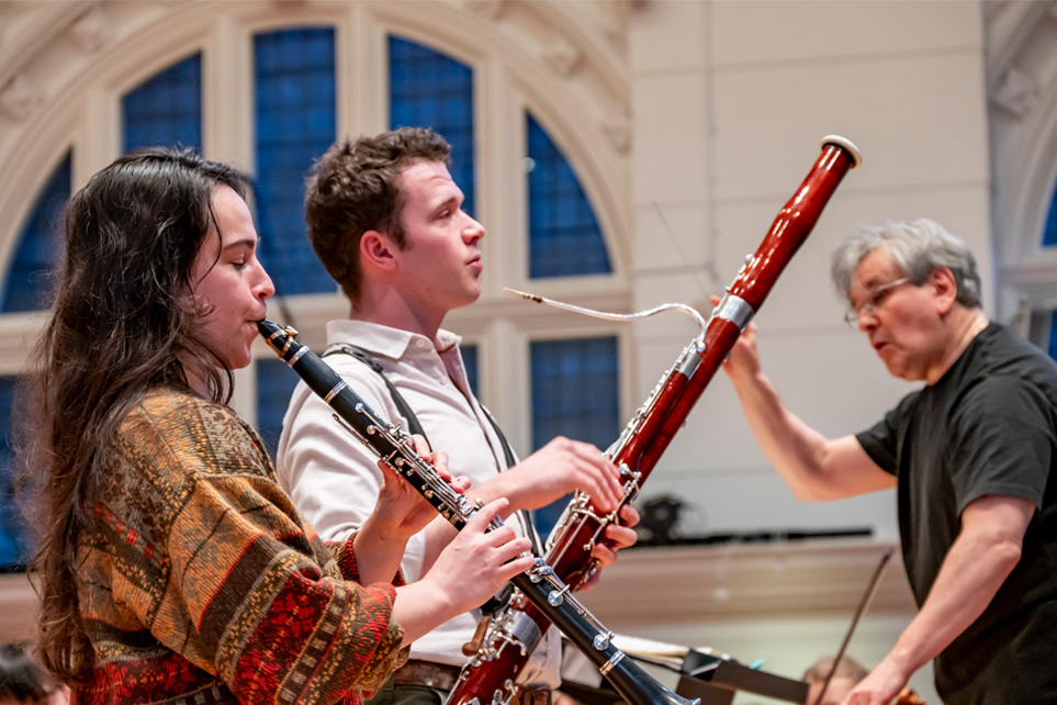 Two students playing the bassoon in a duet in an orchestra rehearsal, standing next to the conductor.